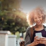 Black single woman smiles in her smartphone while dating online on the go